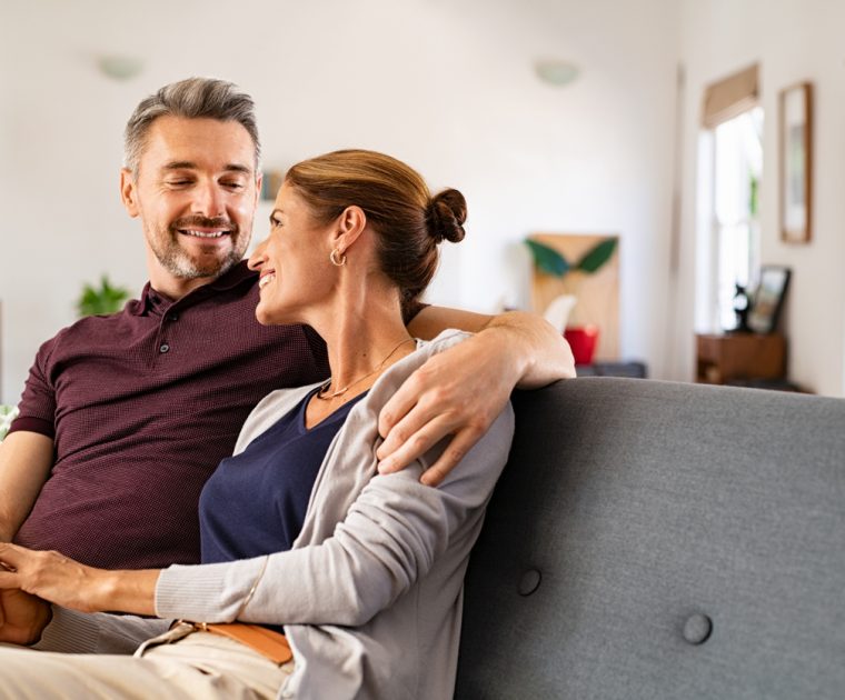 Smiling middle-aged couple sitting at a white table, using a laptop and holding a credit card, planning a purchase or managing finances together.