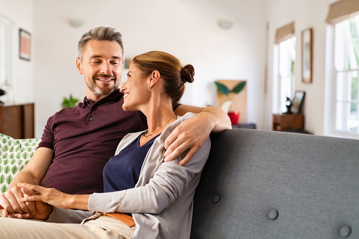 Smiling middle-aged couple sitting at a white table, using a laptop and holding a credit card, planning a purchase or managing finances together.